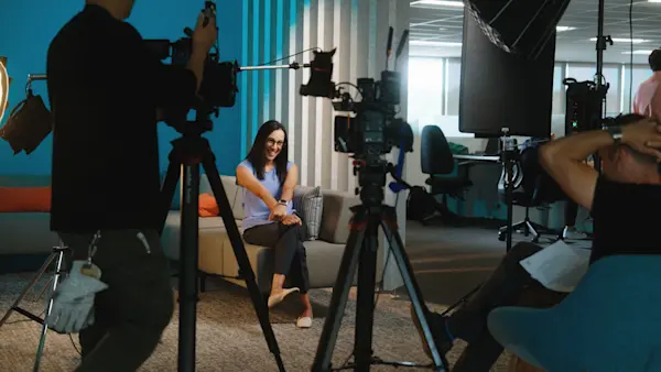 Two cameras frame an interviewee laughing on a sofa in front of a teal wall and slatted panels.