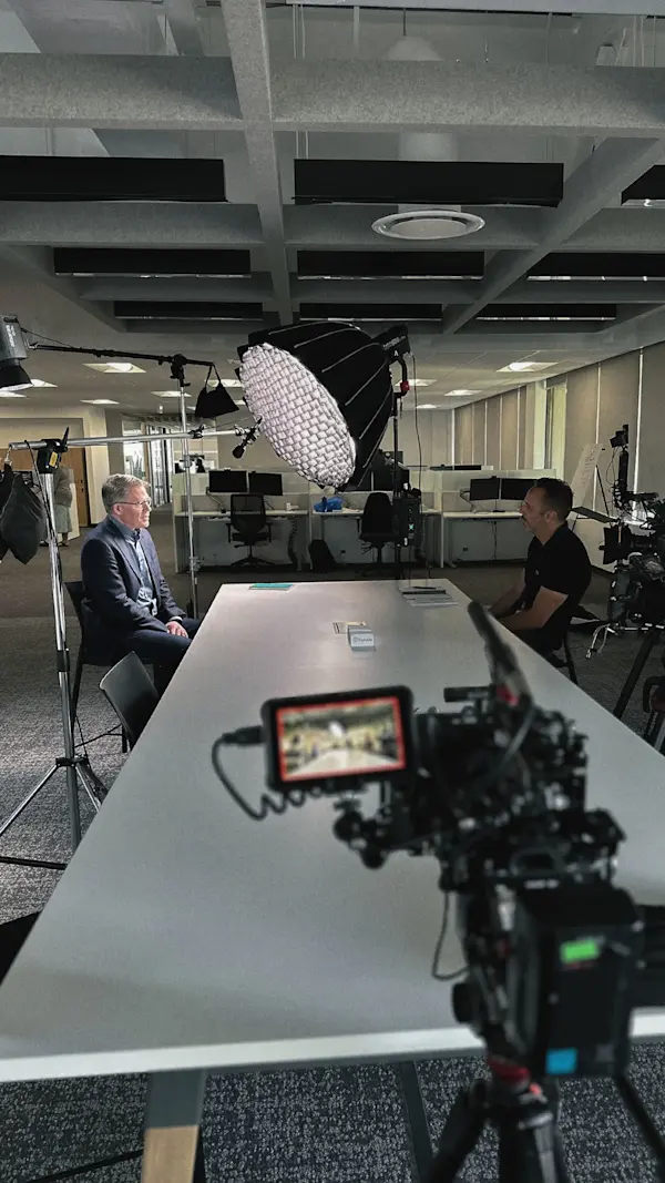 An interviewer and interviewee face each other across a long white table, with a camera monitor up front.