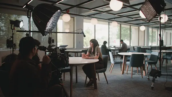 A camera operator films an interviewee holding a coffee cup at a cafe table by the windows.