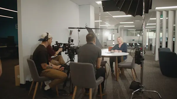Crew in headphones films an interviewee smiling at a round table in a bright office.