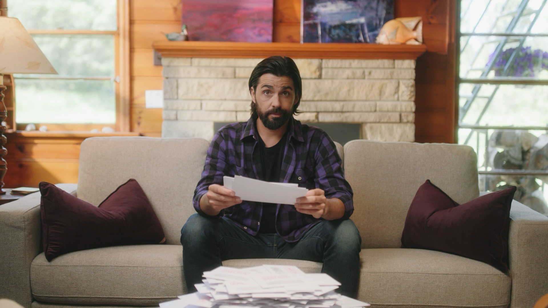 Actor in a purple plaid shirt on a cabin couch behind a pile of bills in Fairstone's Funny Canadian 2 commercial