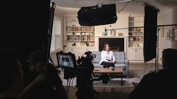 Wide view of a living room interview set with an overhead softbox, black flags, camera and crew facing an advisor