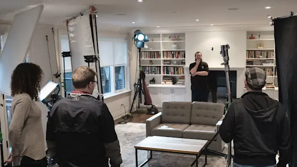 Film crew walking through a living room set surrounded by lights, diffusion frames, and flags before an interview