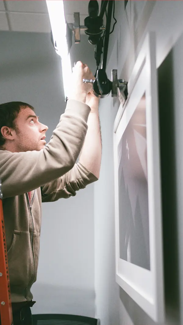 Crew member on a ladder rigging a light above a framed print on an office wall.