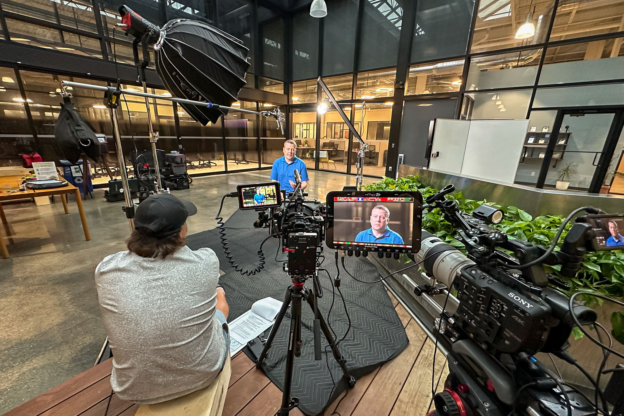 Video crew interviews a man in a blue shirt in an office atrium, with two cinema cameras, a boom mic, and a softbox.