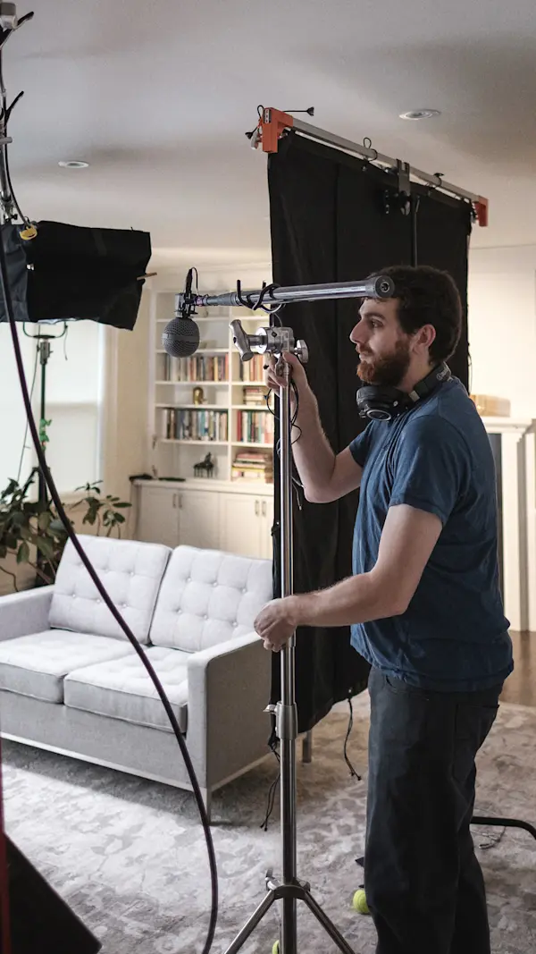Sound recordist adjusting a boom microphone on a stand beside the couch in a living room interview set