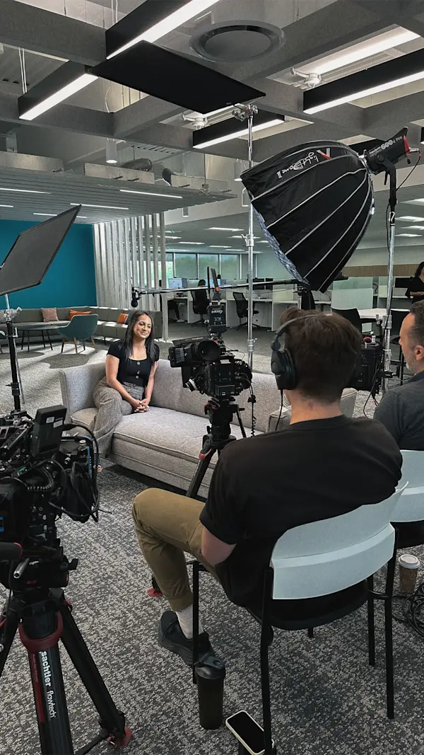 Over the shoulder of a crew member in headphones as an interviewee smiles from a grey sofa.