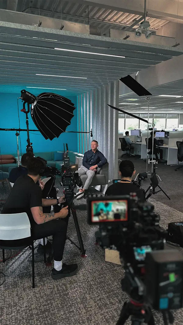 Crew films an interviewee on a grey couch in front of a teal wall, with a camera monitor up front.