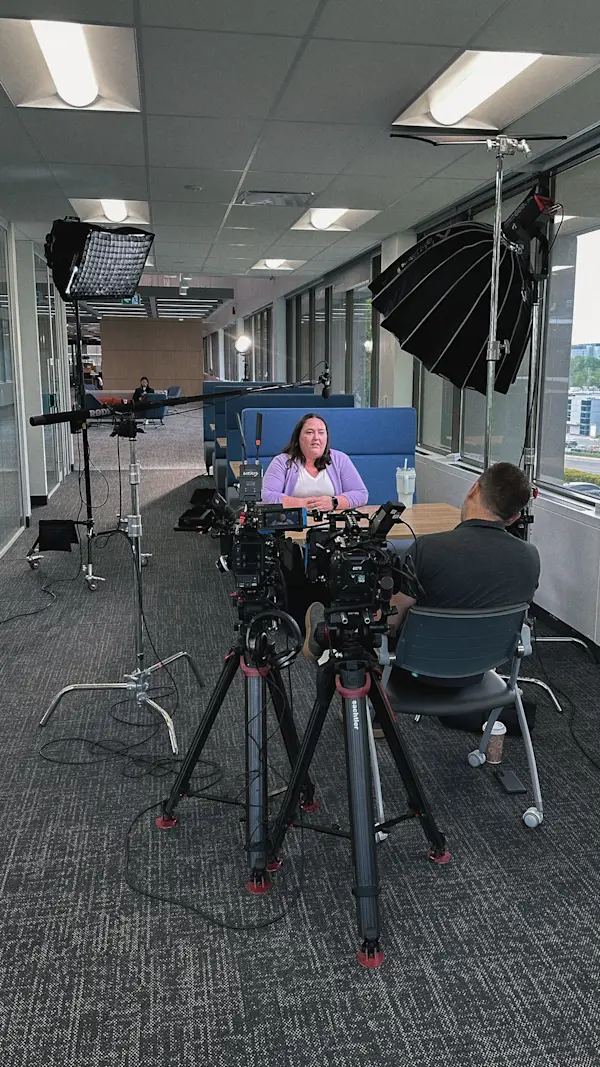 Two cameras on tripods frame an interviewee in a lilac cardigan seated at a blue booth by the windows.