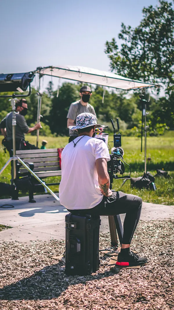 A camera operator sits on a gear case behind the camera, framing a park bench under an overhead silk.