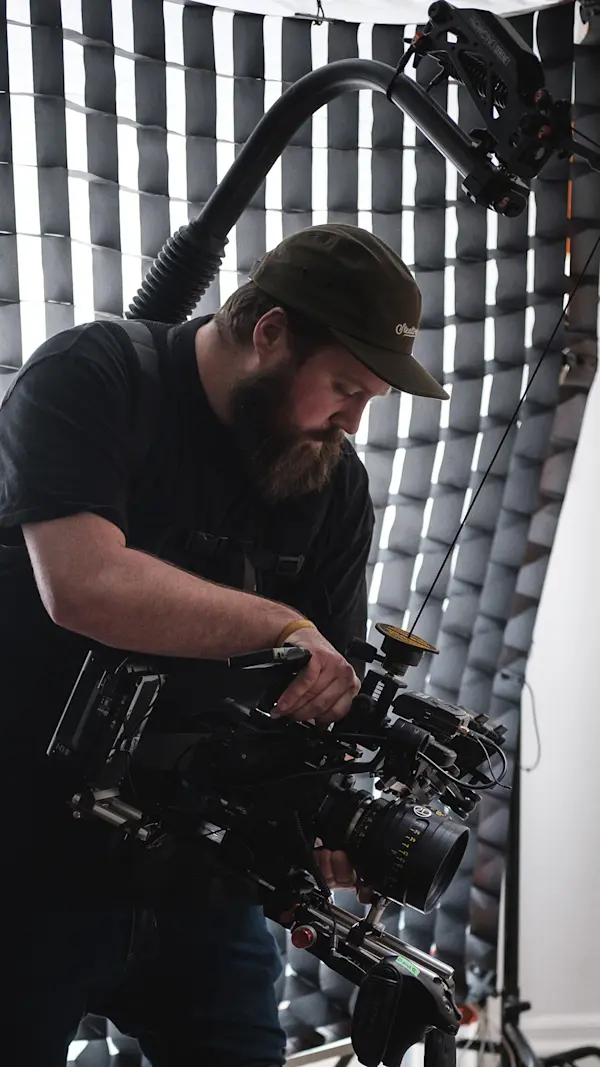 Camera operator checking a cinema camera rig, backlit by a large gridded softbox on the living room set