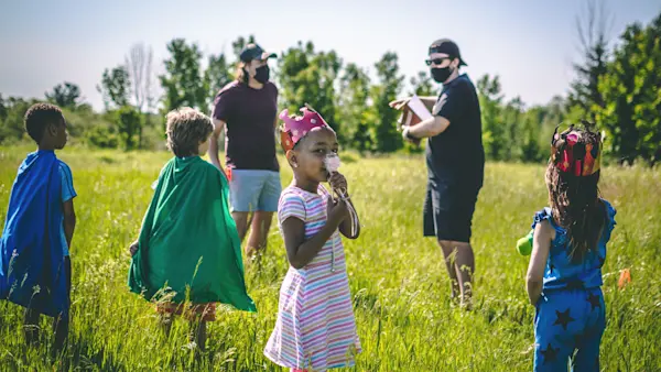 Kids in capes and paper crowns stand in a grassy field while two masked crew members talk behind them.