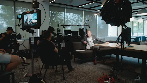 Crew and a camera monitor frame an interviewee leaning back on a dark sofa in a work area.