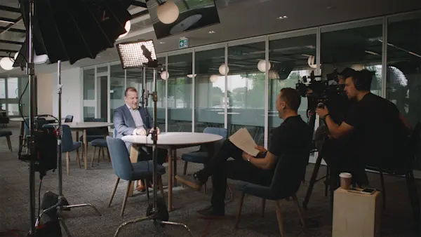 An interviewer reads from notes while a camera operator films the interview at a round table.
