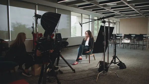 Two crew members sit behind a camera as an interviewee talks in soft light from tall windows.