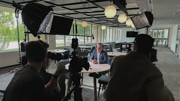 Camera operators frame a seated interviewee in a bright lounge lined with globe pendant lights.