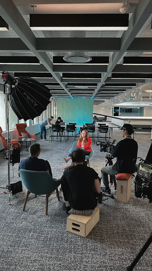 Crew sits on apple boxes around an interviewee in a red blazer in a wide open-concept office.