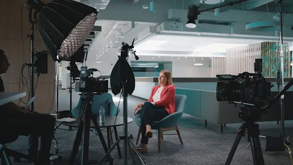 Three cameras surround an interviewee in a red blazer in a modern lounge with teal accents.