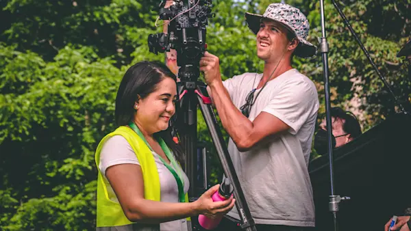 A teacher in a safety vest holds a pink water bottle beside the camera as the operator adjusts the shot.