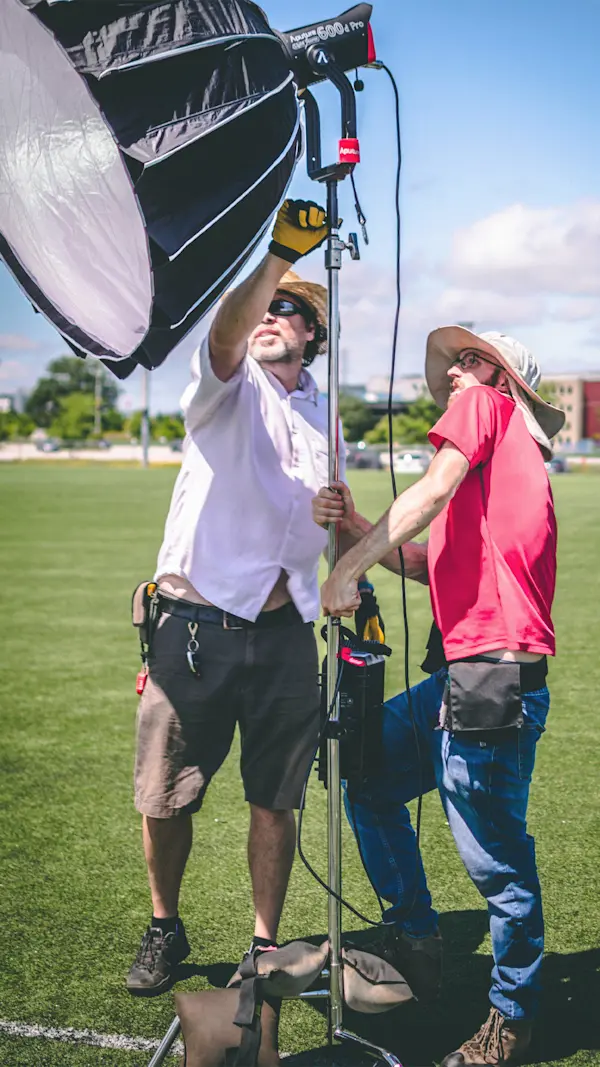 Two crew members in sun hats raise a large softbox light on a turf soccer field.