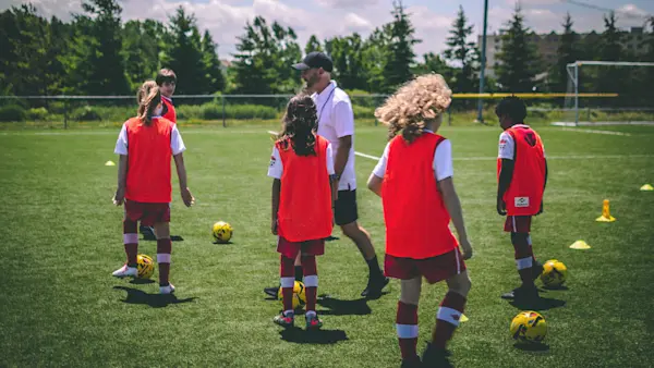 Kids in red pinnies dribble yellow soccer balls around a coach on a turf field.