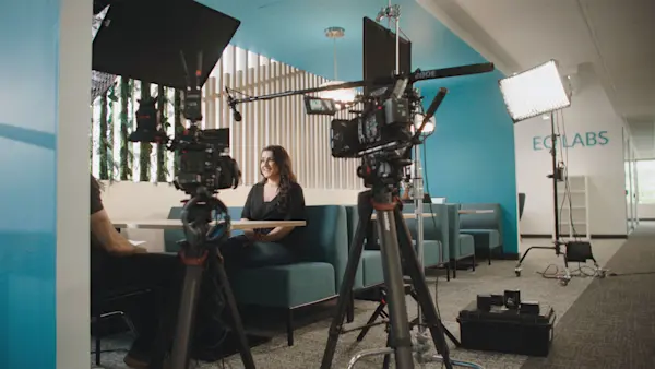 Two cameras and a boom mic frame an interviewee in a teal booth beside the EQ Labs sign.