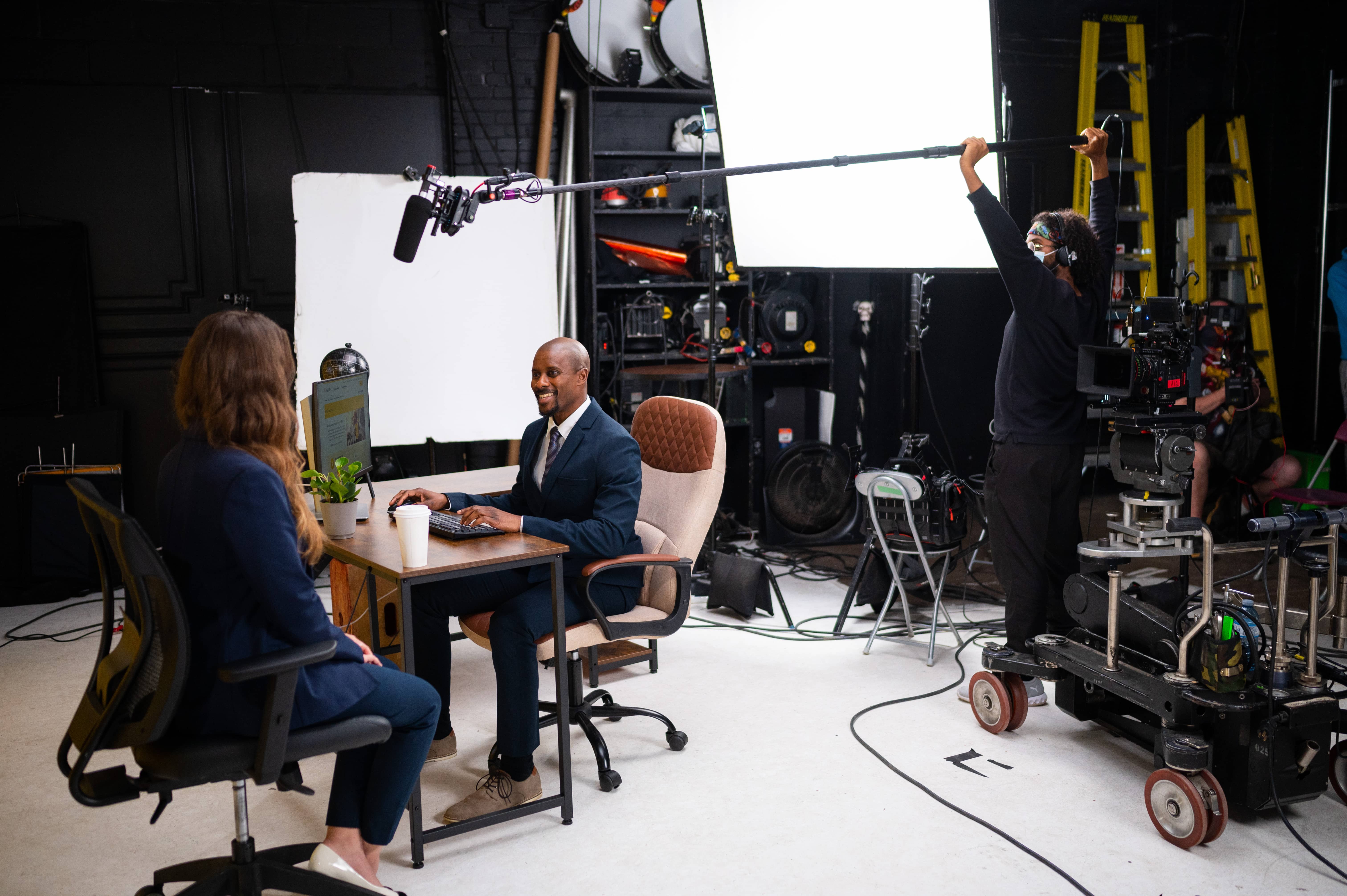 A boom operator and camera crew film two professionals at a desk during a studio video shoot.