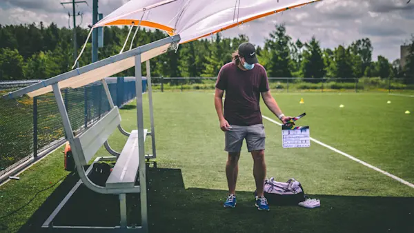 A masked crew member holds a slate beside a soccer team bench, with a pair of white sneakers on the turf.