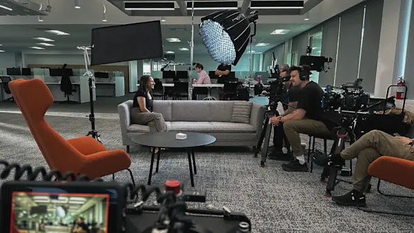 A camera crew films an interviewee on a grey sofa, with a camera monitor in the foreground.