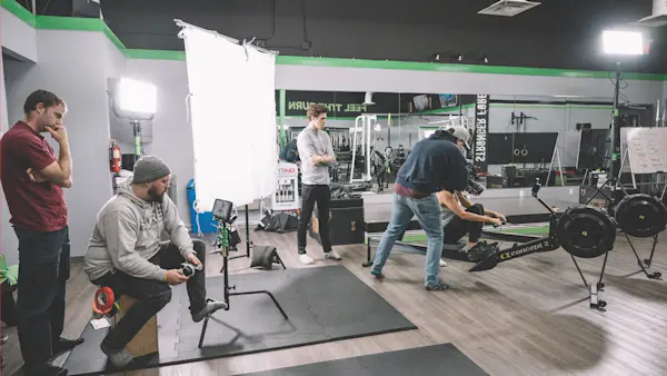 Wide view of the crew filming an actor on a rowing machine in a gym, with lights and a diffusion frame.