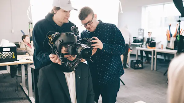 Two crew members fitting a POV helmet camera rig onto a seated actor in a suit.