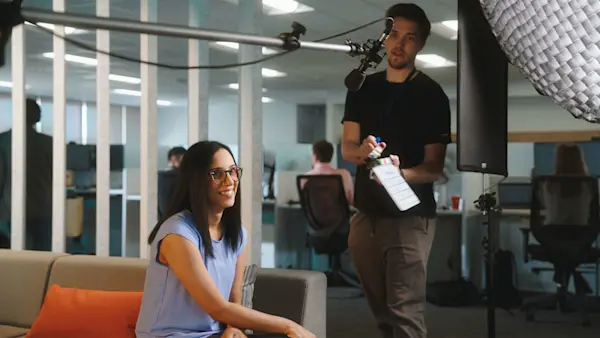 A boom operator holds a slate and mic over a smiling interviewee seated on a sofa.