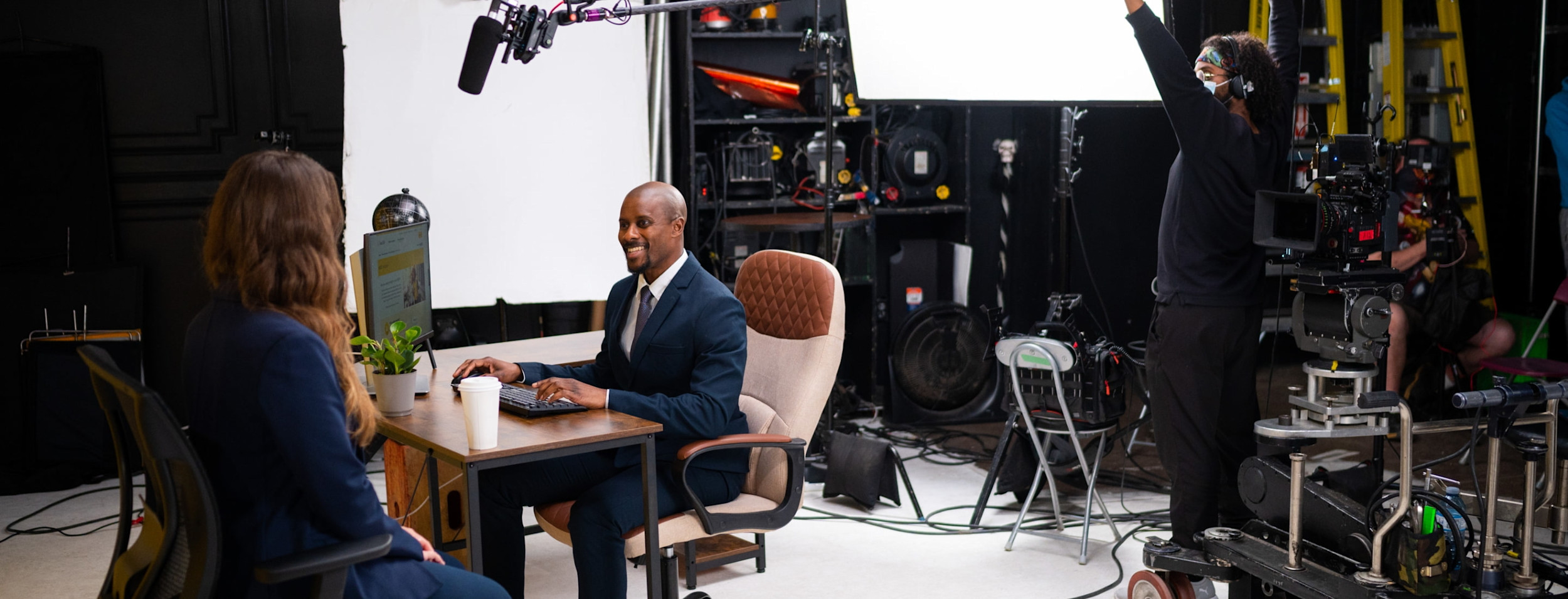 A boom operator and camera crew film two professionals at a desk during a studio video shoot.
