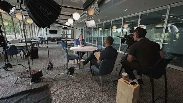Two-camera crew films an interview at a round table, with softbox lights and a producer holding notes.