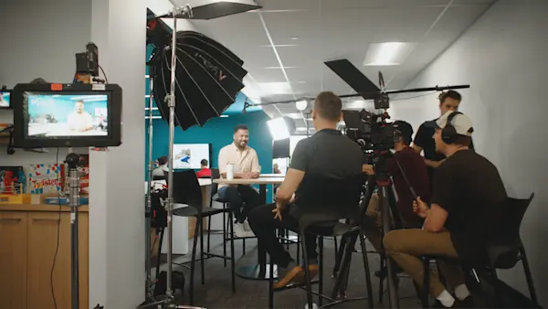 Full crew sits in on an interview in a teal room, with the shot showing on a monitor at left.
