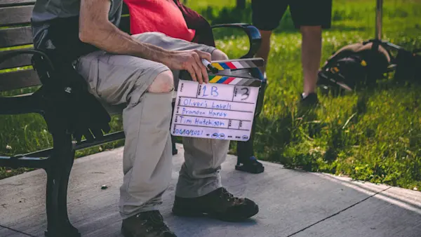 A crew member sits on a park bench holding a slate marked Oliver’s Labels, scene 1B, take 3.