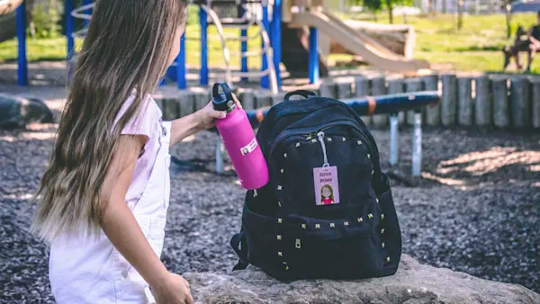 A girl at a playground puts a pink water bottle with a name label into a backpack with a matching name tag.