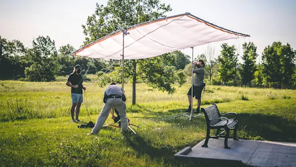 Three masked crew members rig a large white overhead silk above a park bench in an open field.