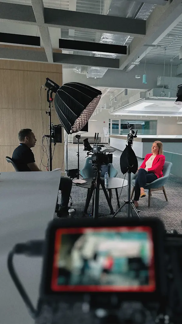 Seen past a blurred camera monitor, an interviewer talks with an interviewee in a red blazer.