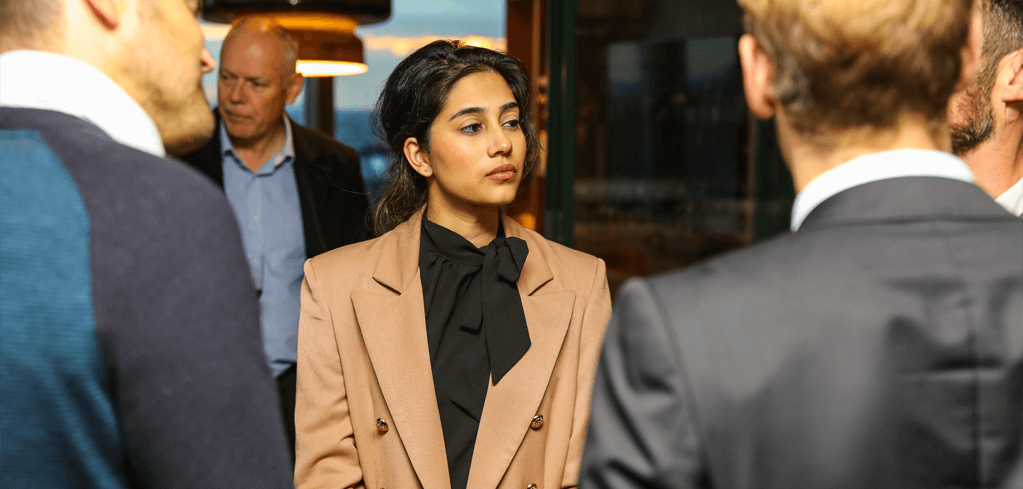 Woman in beige blazer and black bow tie standing among businesspeople at a professional gathering.