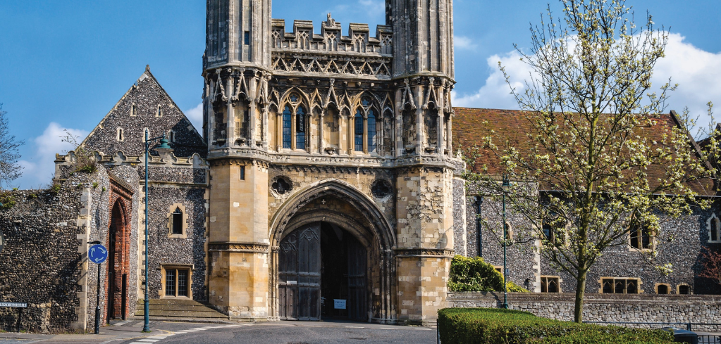 A Gothic stone cathedral gatehouse with ornate arched entrance, flint walls, and a tower under a bright blue sky.