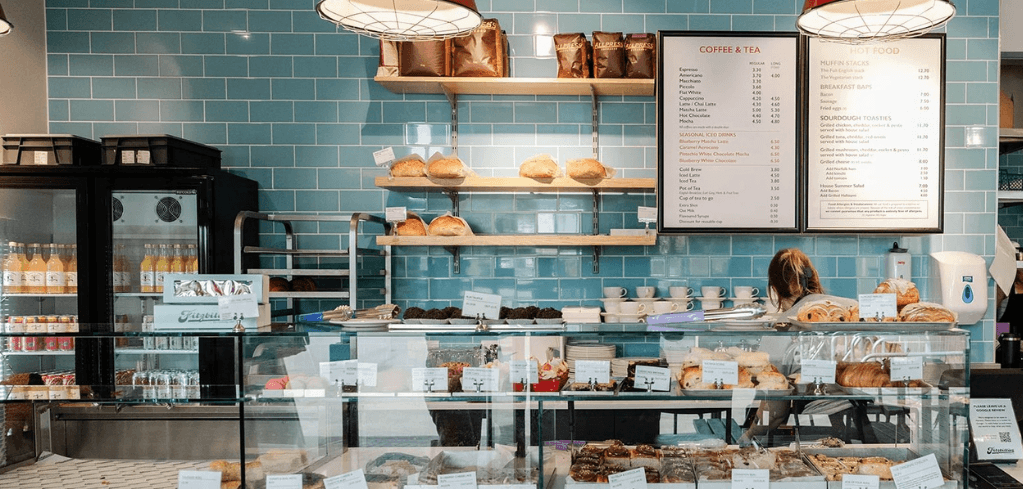 A bright café counter with a glass display case of pastries, blue tile walls, shelves of bread, and menu boards above.