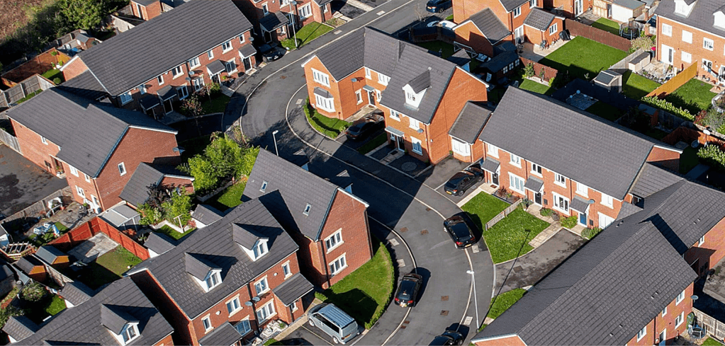 Aerial view of a residential neighborhood with red brick houses, gray roofs, small yards, and curved streets.