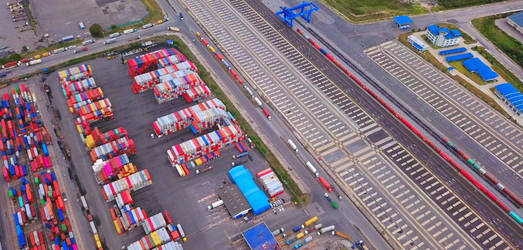 Aerial view of a shipping container yard with colorful stacked containers next to rail tracks and loading facilities.