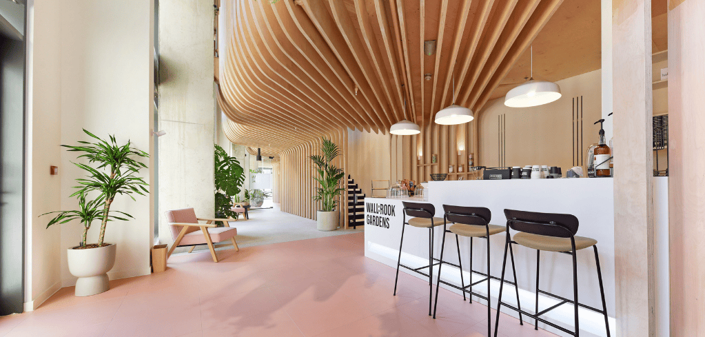 Modern café with curved wooden ceiling, pink floor, white counter, bar stools, and potted plants.