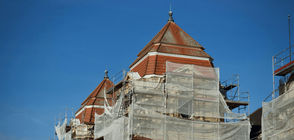 Historic building with red-tiled pyramid rooftops under renovation, surrounded by white scaffolding and netting against a clear blue sky.