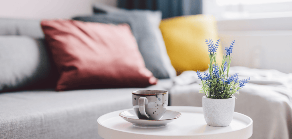Cozy living room with coffee cup and lavender plant on white table, colorful pillows on sofa in background.