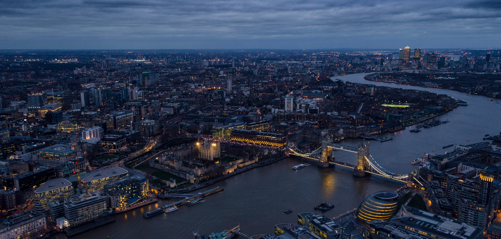 Aerial night view of London with Tower Bridge illuminated over the Thames River, city lights stretching to the horizon.