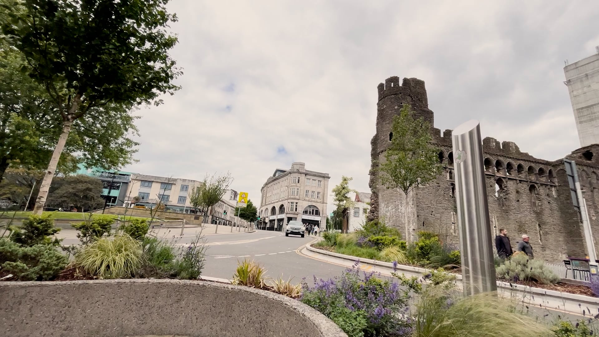 Medieval stone castle ruins alongside modern buildings in a town center with landscaped gardens and curved road.