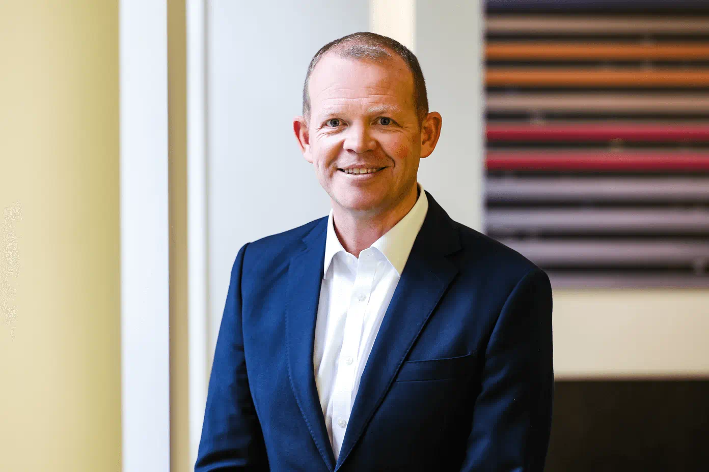 A person in a navy blue suit and white shirt smiling at the camera in a professional office setting.